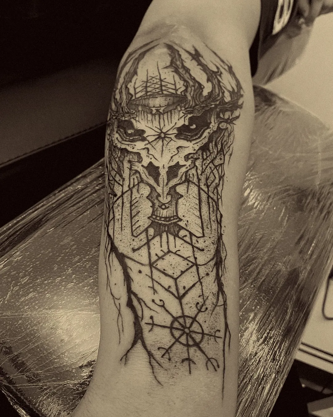 Calavera con corona de espinas sobre runas y un vegvísir, en blackwork en el antebrazo