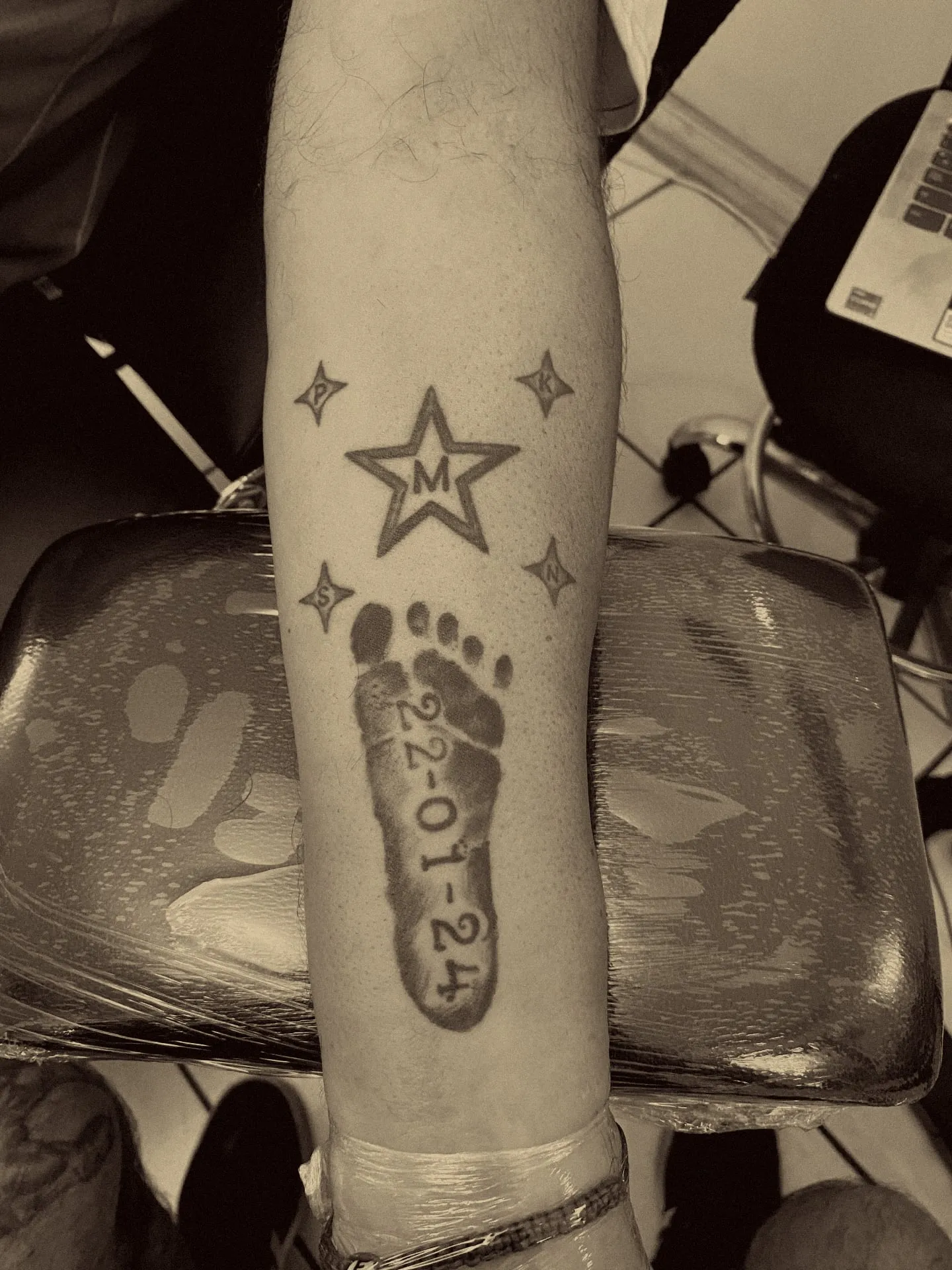 Estrella con inicial, estrellas pequeñas y huella de bebé con fecha, cover-up a mano alzada en el antebrazo
