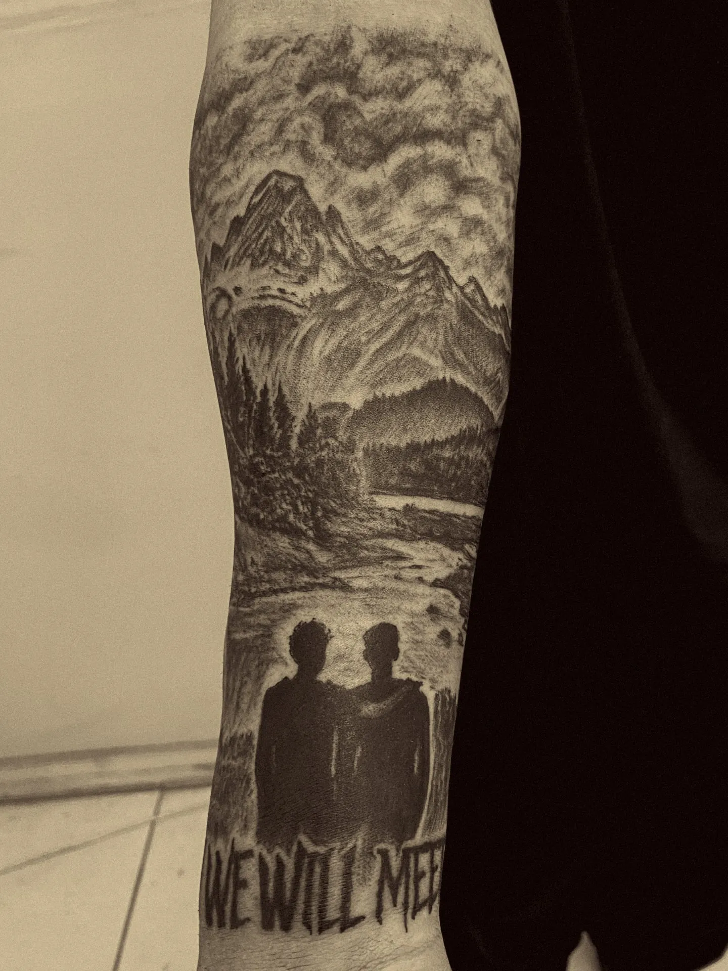 Montañas, bosque y río con dos siluetas abrazadas y el texto We will meet again, en negro y gris en el antebrazo