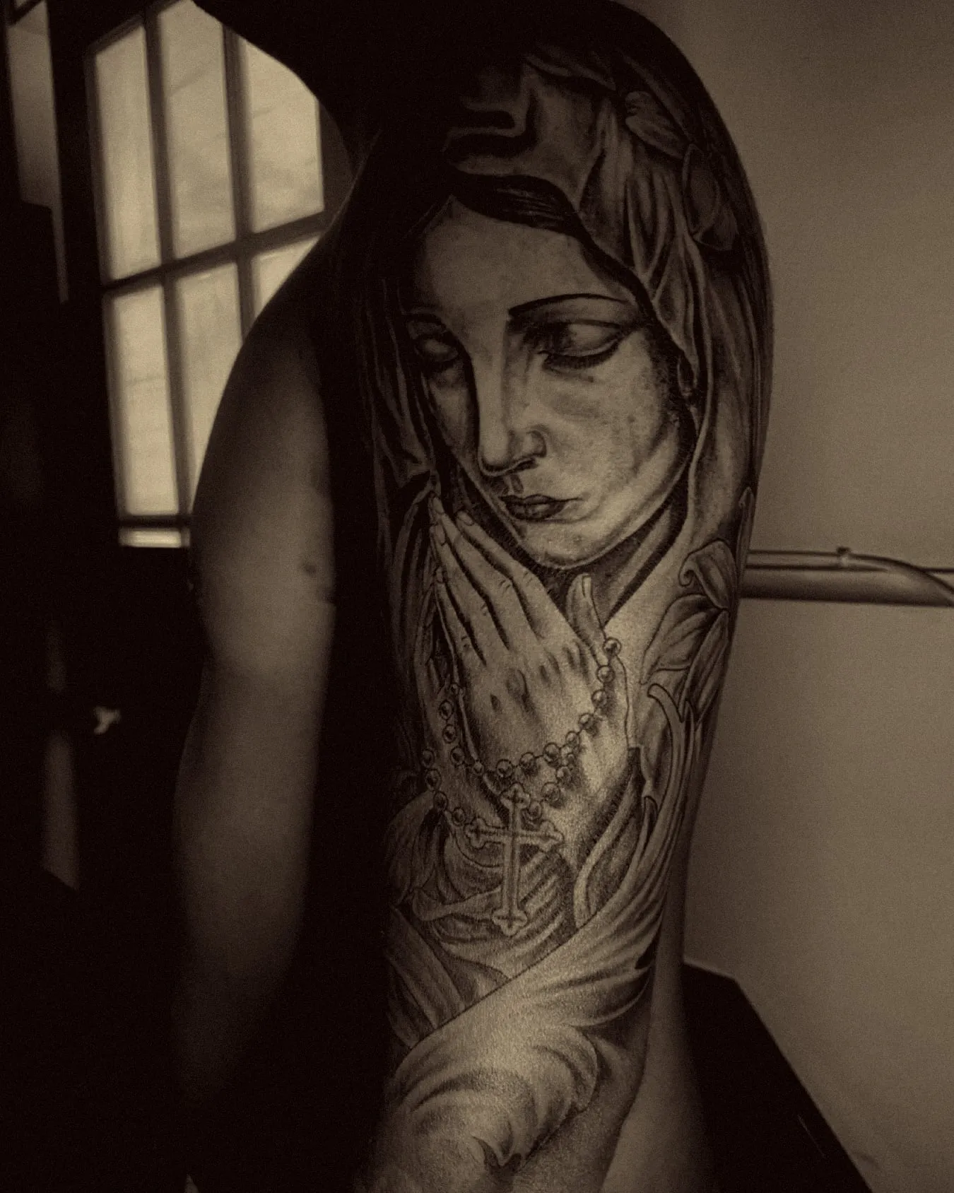 Virgen con las manos juntas y un rosario, en realismo negro y gris en el brazo